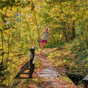Trailrunnerin läuft auf einem herbstlichen Waldpfad über eine kleine Holzbrücke bergab